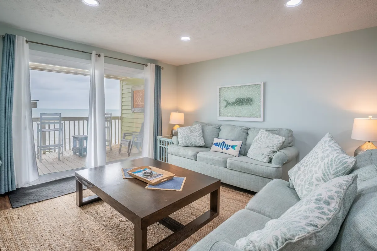 The image showcases a bright, inviting living area with plush sofas arranged around a coffee table adorned with books, all facing a sweeping window that frames breathtaking ocean views. Sunlight pours in, illuminating the relaxing space and drawing attention to the balcony where guests can greet the sunrise over the Atlantic. The scene radiates warmth, comfort, and the promise of peaceful moments shared with family or friends.