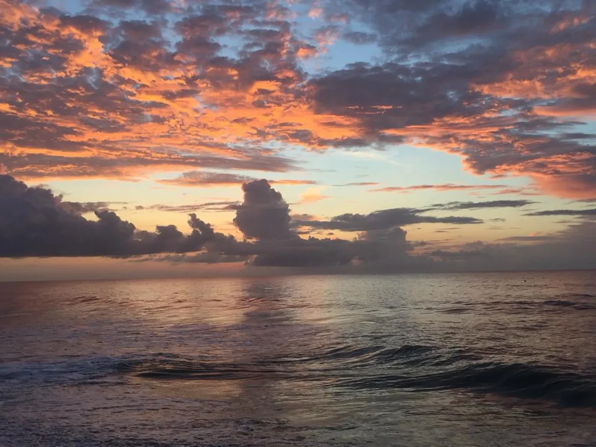 A dramatic view of the ocean at sunset, with waves rolling in and clouds painted orange and purple by the setting sun. The mood is peaceful and romantic, ideal for a reflective walk or sharing a special moment along the shore.