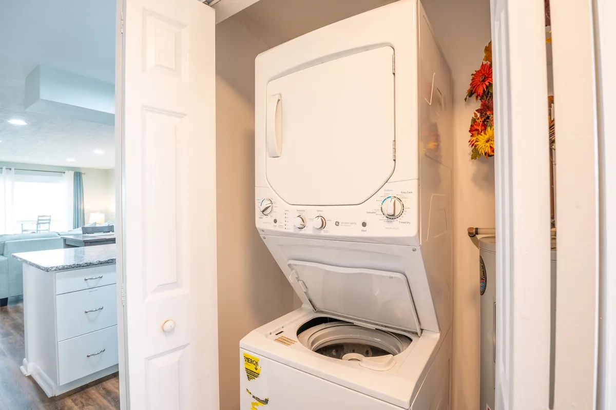 A neatly organized laundry closet houses a stacked washer and dryer, enhanced by decorative touches like flowers for a welcoming, home-like feel. The setup emphasizes practicality and convenience for guests.