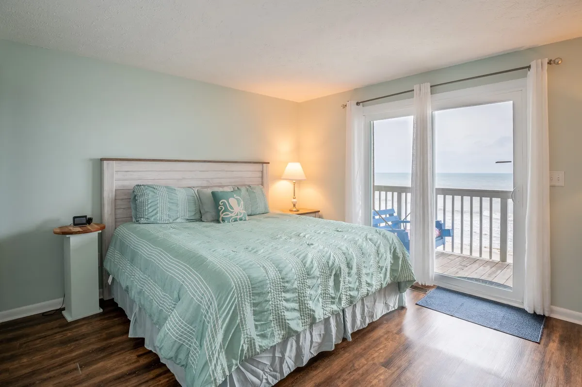 A king-sized bed adorned with teal bedding is the primary focus, complemented by a bedside lamp and a sliding glass door that opens to a private balcony, allowing natural light to flood the serene space. The inviting setup hints at a tranquil retreat with direct access to ocean views and fresh air.