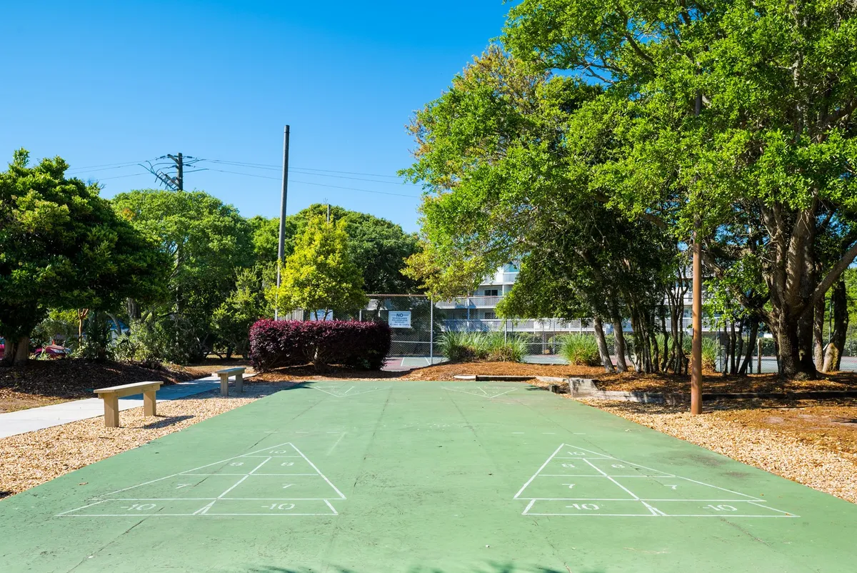A dedicated outdoor shuffleboard court framed by benches and lush trees, inviting families for friendly competition or relaxed spectating. The court is spacious and accessible, set in a peaceful, green environment that encourages social interaction and outdoor play.
