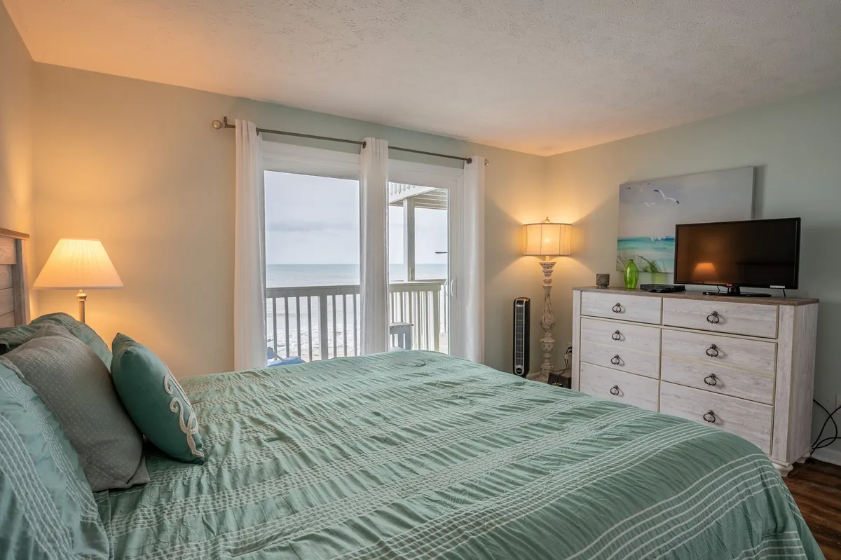 A tranquil bedroom scene featuring a bed with teal bedding, a large window overlooking the ocean, a bedside lamp, and a dresser with television. The space feels inviting and restful, suggesting comfort and essential touches like fresh linens.