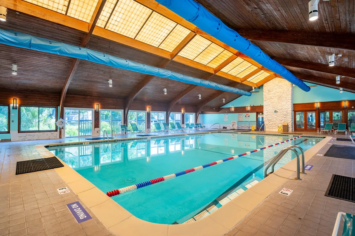 A bright indoor swimming pool area with wooden ceiling beams, large windows letting in natural light, lounging chairs along the poolside, and an overall airy and refreshing ambiance. Ideal for year-round enjoyment.
