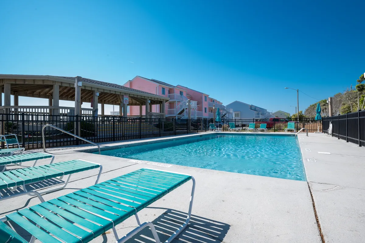 An outdoor swimming pool surrounded by inviting lounge chairs and nearby buildings, set under a bright, open sky. The pool area is well-kept, offering plenty of space for families to swim, relax, or gather for social activities. The presence of sun umbrellas and ample seating creates a lively and comfortable atmosphere suitable for both play and relaxation.