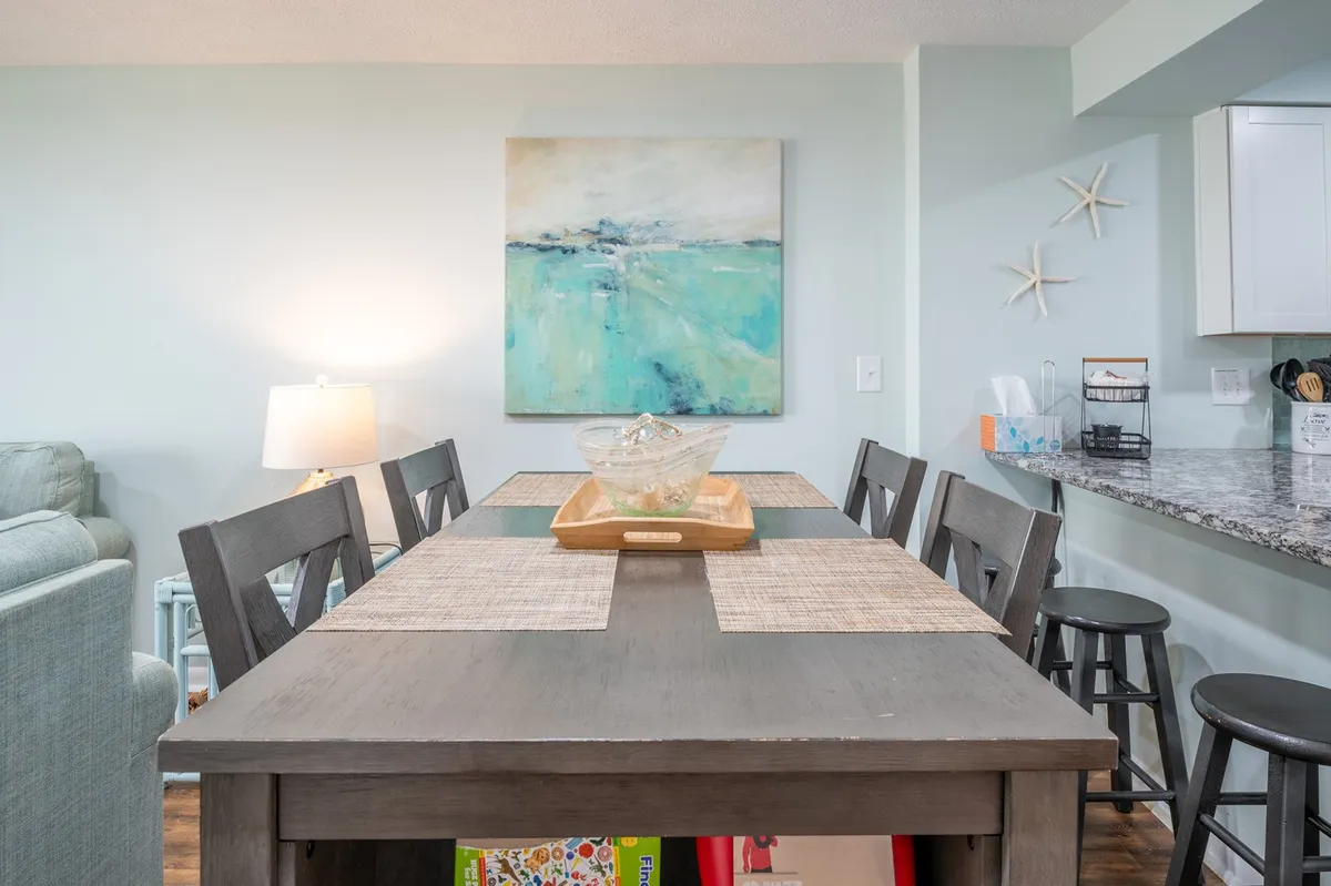 A dining area with a contemporary table, art on the walls, and a lamp providing warm illumination. The kitchen counter is visible, tying together the living and dining spaces for a sense of continuity.