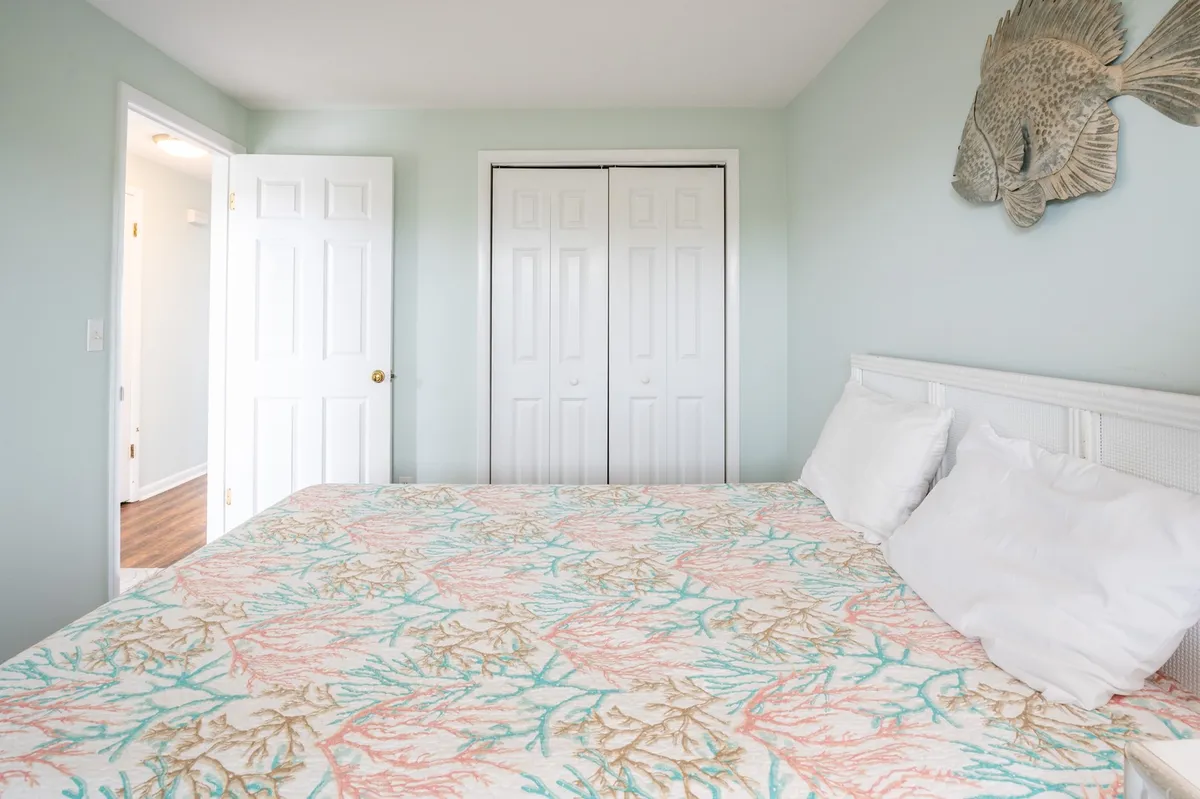 A cozy queen bedroom features coral-patterned bedding, fish-inspired wall decor, and large double closet doors. Soft lighting and a neutral palette create a restful and welcoming environment, ideal for guests seeking comfort and privacy.