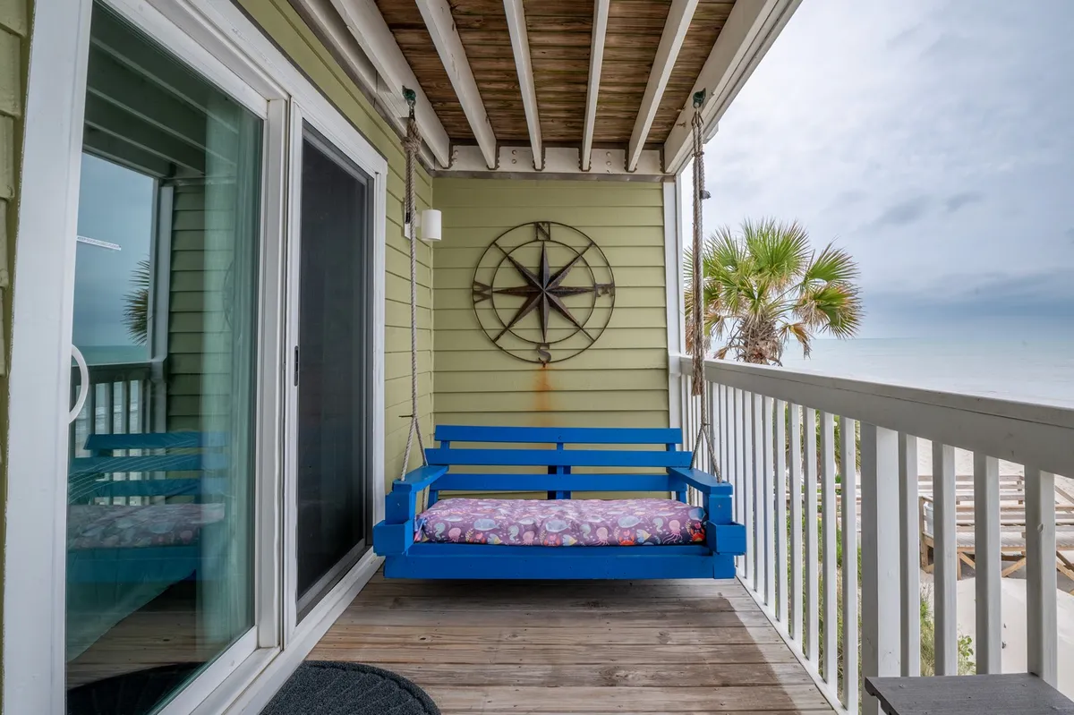 A private deck with a blue porch swing, star compass wall art, ocean views, and palm trees. The deck is a tranquil spot for enjoying sunrises, sipping coffee, or relaxing with the sea breeze.