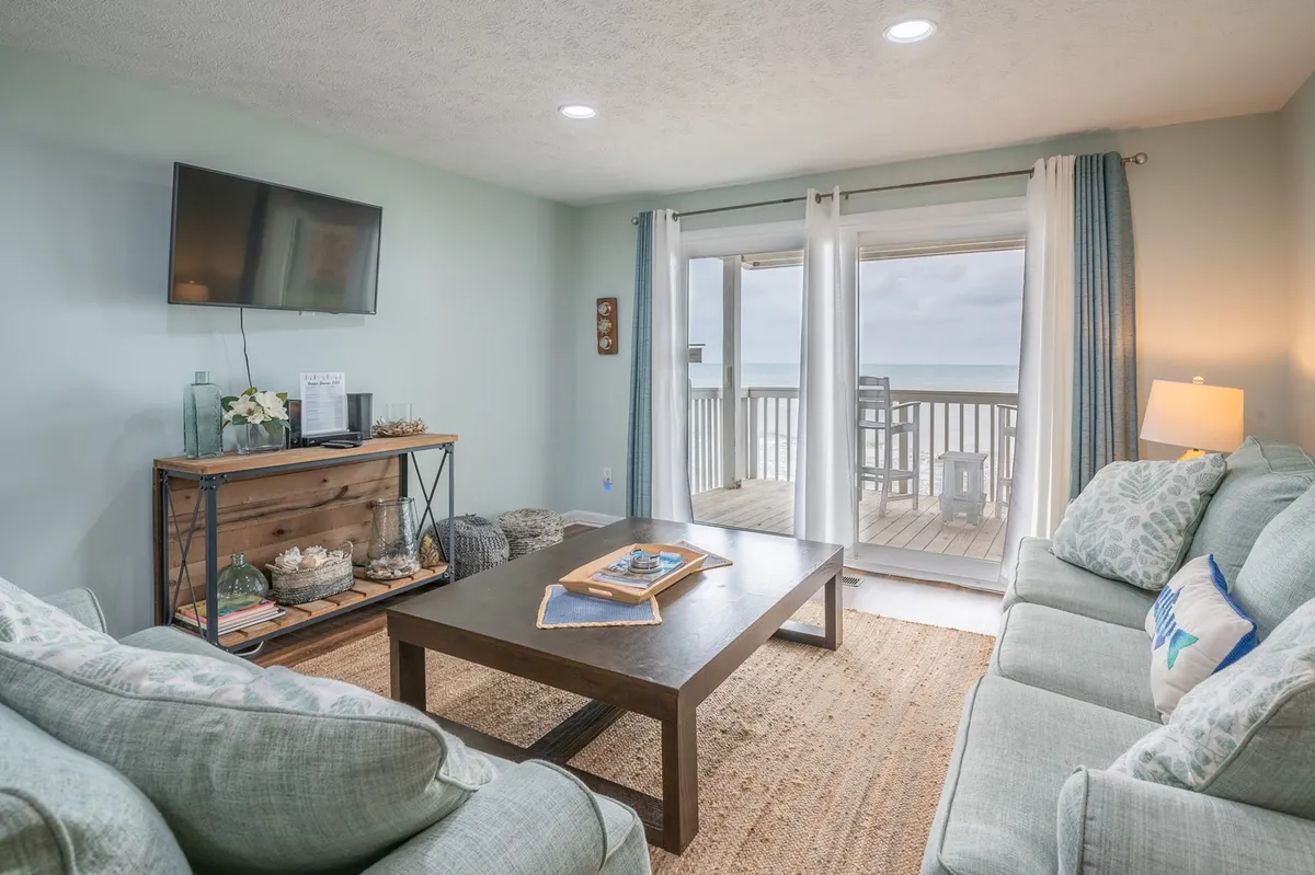 This image features a cozy living room with comfortable sofas arranged around a coffee table, a smart TV mounted on the wall, and large windows leading out to a balcony. The room is bright and welcoming, with the balcony hinting at the nearby ocean beyond the glass doors.