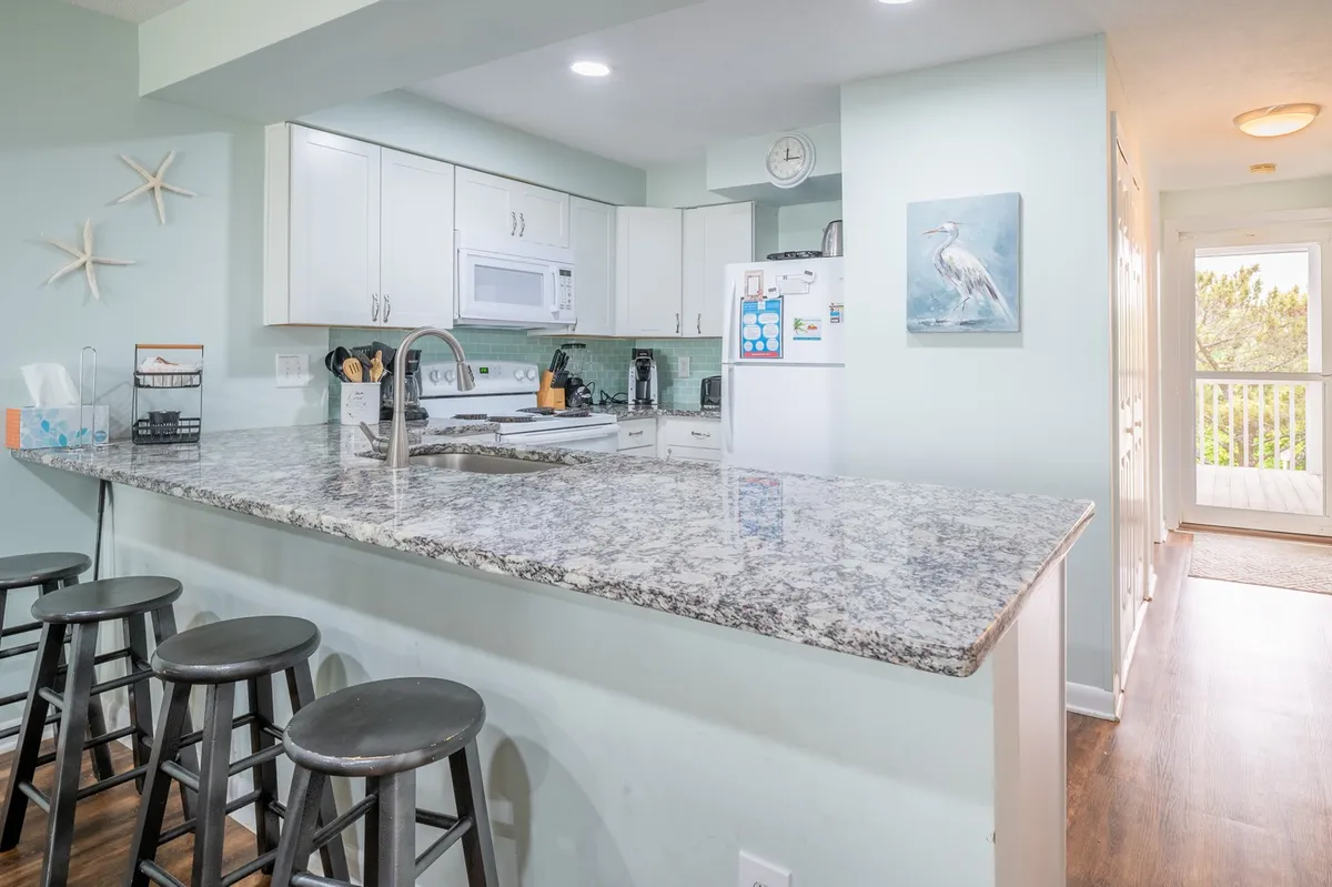 A modern kitchen with granite countertops, white cabinetry, sleek appliances, and comfortable island stools. Artwork adds a personal touch to the cheerful, functional space, ideal for preparing meals or gathering for conversation.
